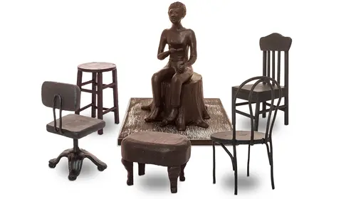 A miniature, or maquette, of a sculpture in reddish-brown clay depicting Lorraine Hansberry sitting on a tree stump surrounded by five different kinds of chairs