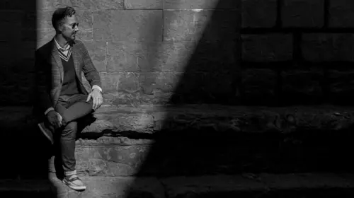 Monochrome photo of Joey Stocks sitting on a stone bench in front of a stone wall illuminated by a triangular slice of light